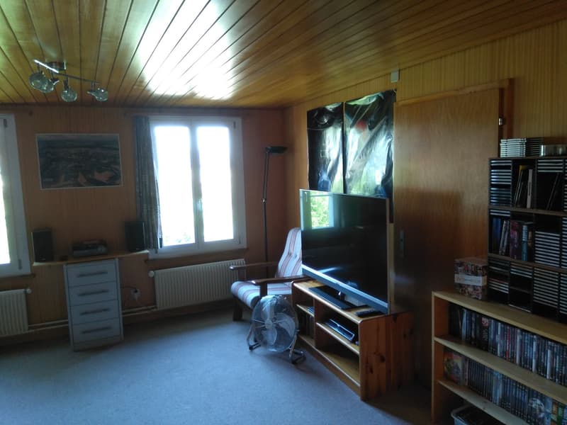 Living room with wooden floor, wall-mounted radiators, desk, television, couch, bookshelf with media, fan, artwork on wall.