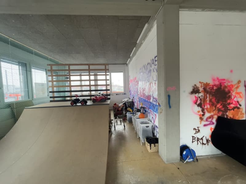 Industrial style room, large wooden ramp, graffiti on the walls, windows with metal grills, trolleys and items stored, large pillars.