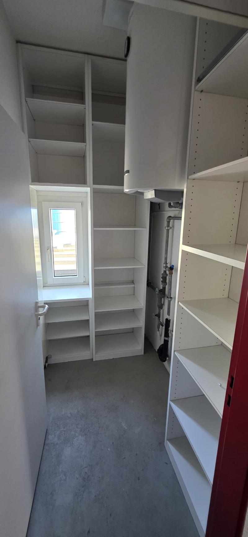 walk in closet with white shelves, small window, door on the left, pipes behind the closet