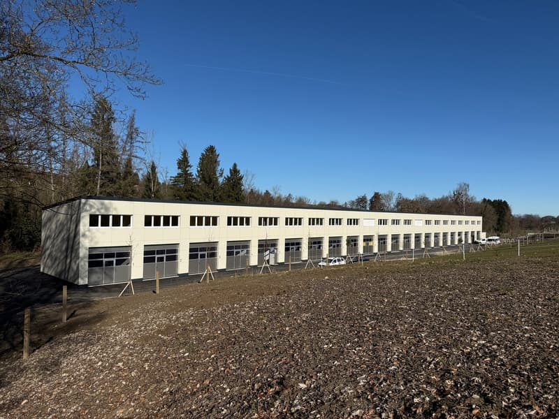 A long, white building with multiple windows and garage doors, surrounded by a field and trees, with cars parked in front.