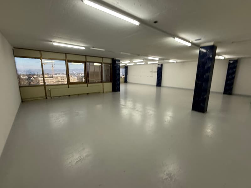 Empty room, white walls, white floor, large windows, ceiling with light fixtures, exterior view of buildings