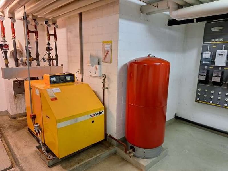 Yellow gas boiler, red water tank, pipes, electrical panels, white wall, concrete floor.