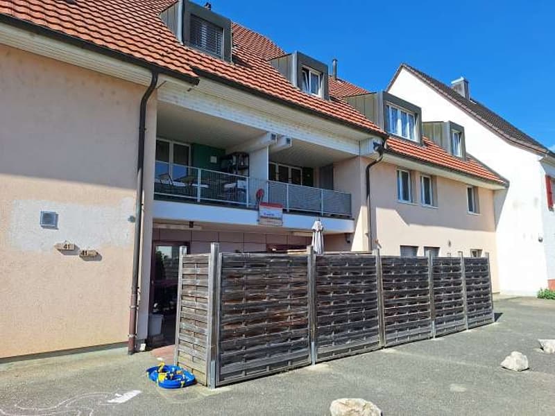 Two-story building with brown roof, beige walls, glass windows, balcony with metal railings, wooden fence at the entrance