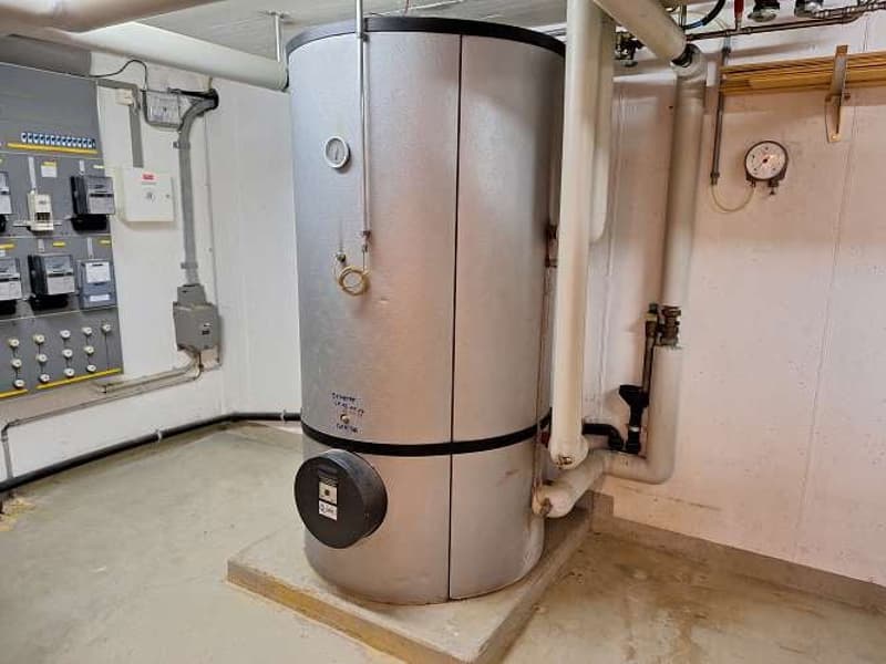 Water heater with gauges and pipes, electric panel with meters and breakers, concrete floor.