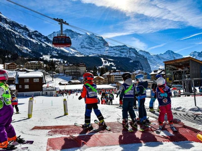 Skiers and snowboarders are present at a snowy mountain area with mountains and snow-capped mountains in the background, there are two gondola cable cars in the foreground.