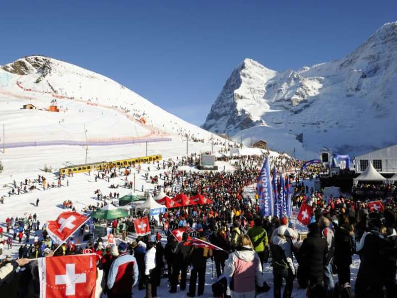 A large crowd gathered at a ski event in snowy mountains with flags and tents