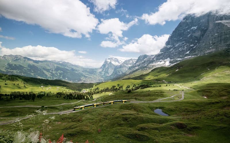 Mountain valley with lush green hills and a yellow and green train on the tracks under a partly cloudy blue sky
