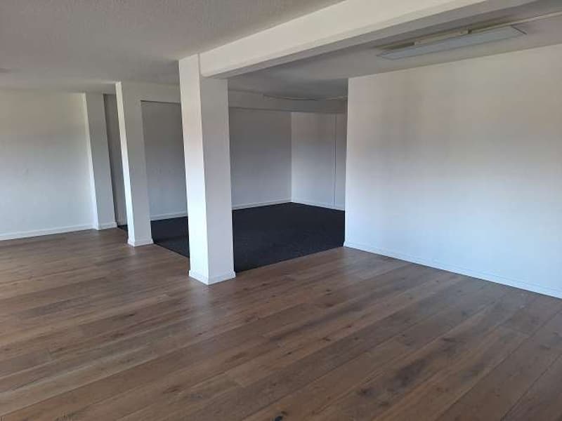 The room has a rectangular shape with wooden floors, white walls, and black carpeting. There are columns in the middle of the room and two openings leading to another room.