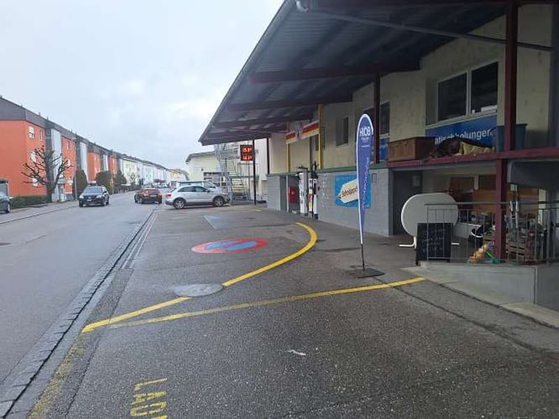 residential area, gray parking lot with yellow lines and 'P' sign, building with metal awning and storage boxes on second floor, red car and sedan driving by