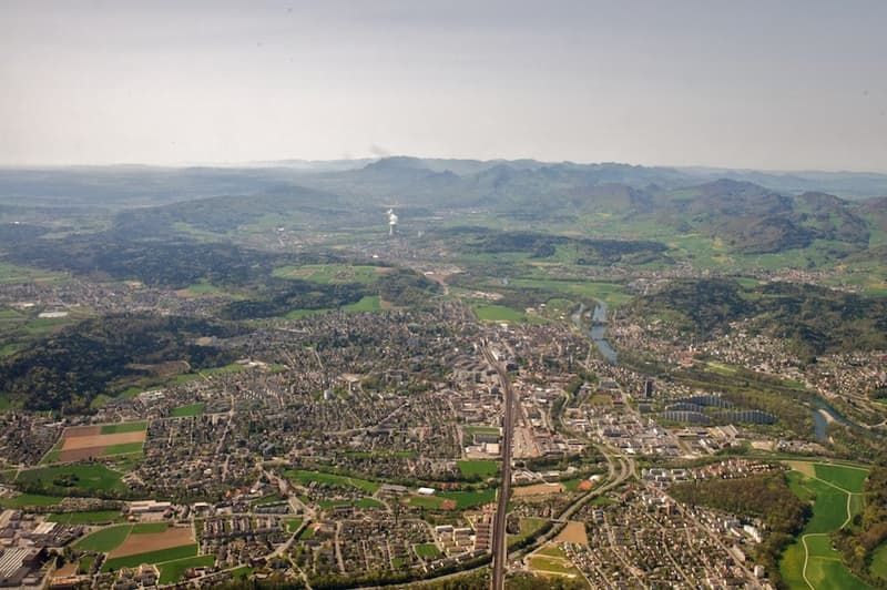 Aerial view of city surrounded by mountains, river, buildings, green fields, infrastructure
