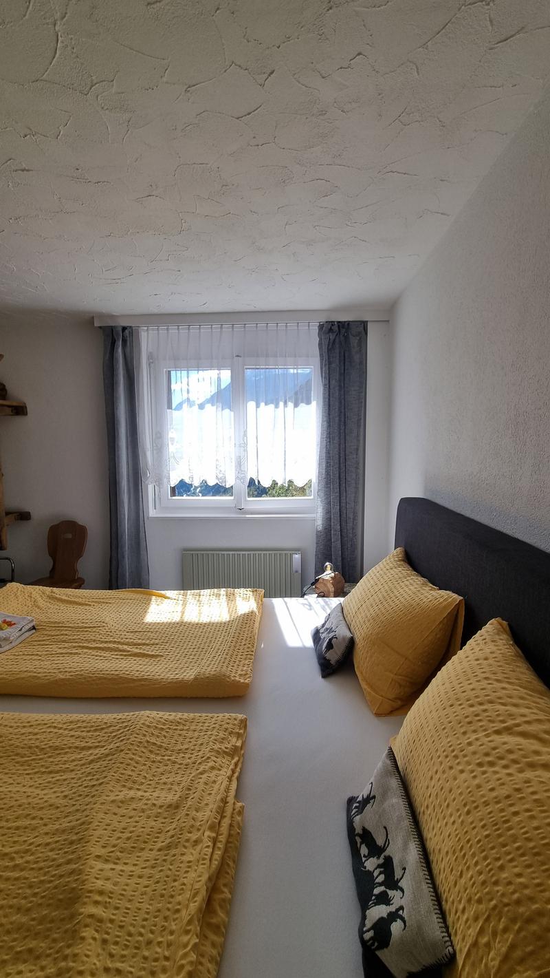 Bedroom with two beds, a chair, a desk with books, a window with curtains, and a view of the outside.
