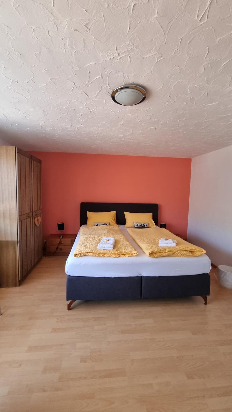 A double bed, night stands, and a closet are in the room. The bed has two yellow pillows and two yellow duvets. The nightstands have lamps and small tables with white towels on them. The wall is painted in orange and white. The closet is wooden and mounte
