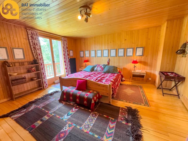 Cozy wooden bedroom with large windows, wooden paneled walls, a double bed with colorful bedding, side tables, lamps, and framed artwork on the walls. The room also has a balcony or terrace visible through the windows.