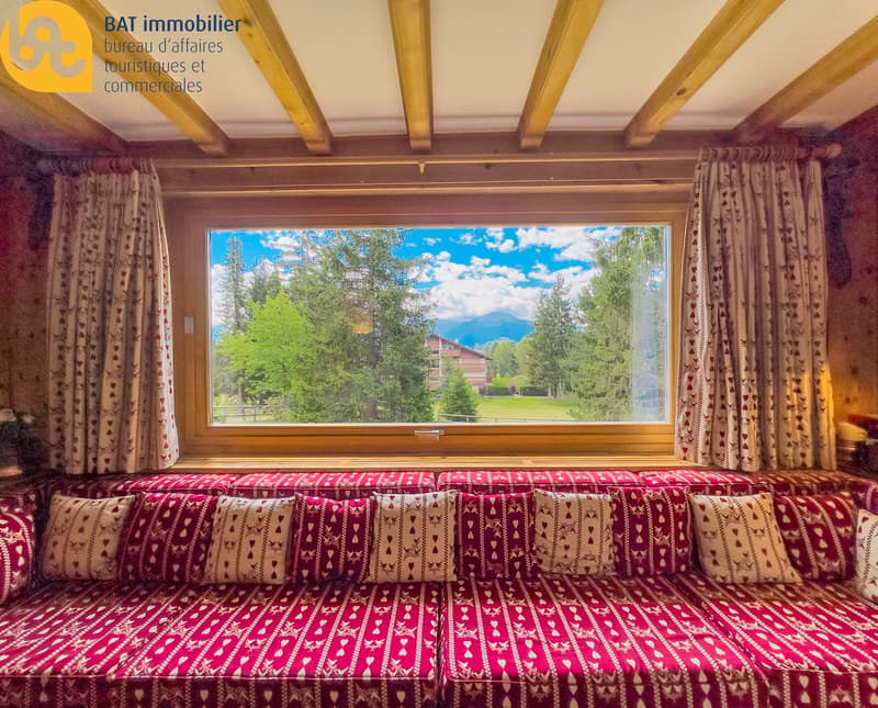 Large window with scenic view of trees and mountains, wooden ceiling beams, patterned curtains and cushions on a couch