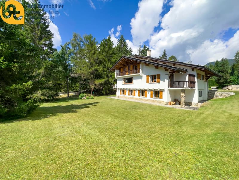 2-story chalet-style house with white exterior, wooden balconies, and large windows. The house is surrounded by a well-maintained lawn and trees, providing a scenic, natural setting.