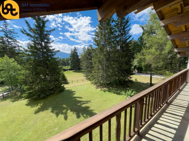 Wooden chalet-style building with a balcony overlooking a large grassy area surrounded by pine trees and a clear blue sky with fluffy white clouds.
