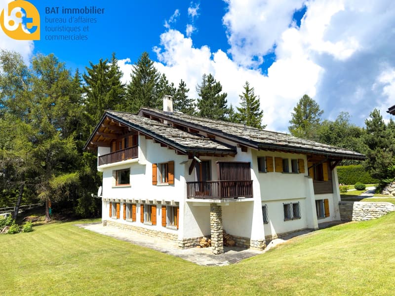2 story chalet-style house, white exterior with wooden accents, stone foundation, slanted roof with wooden beams, balcony, surrounded by pine trees and green lawn