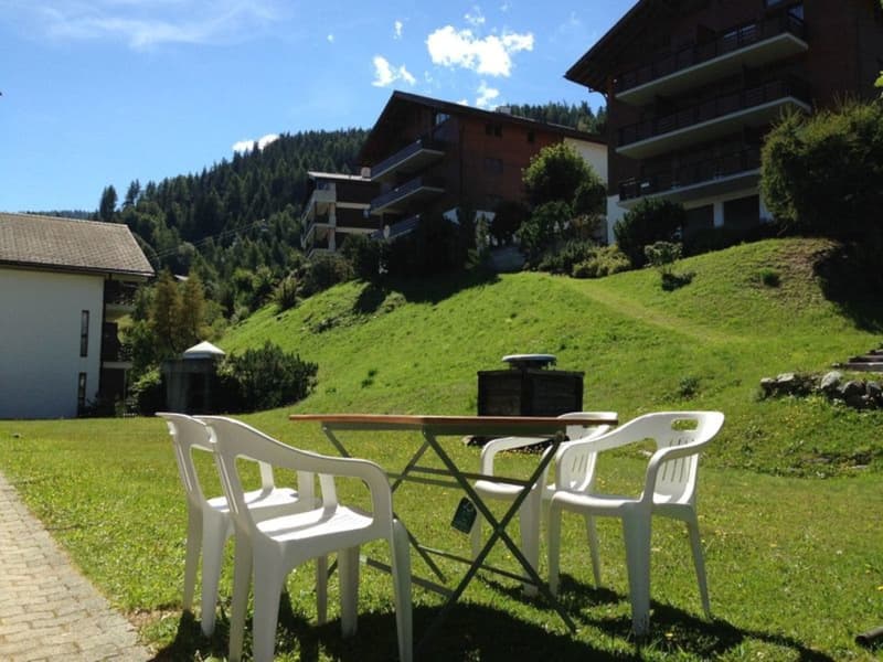 The image shows an outdoor seating area with a small table and chairs set up on a grassy hill. The area is surrounded by a forested mountainous landscape with several buildings visible in the background.