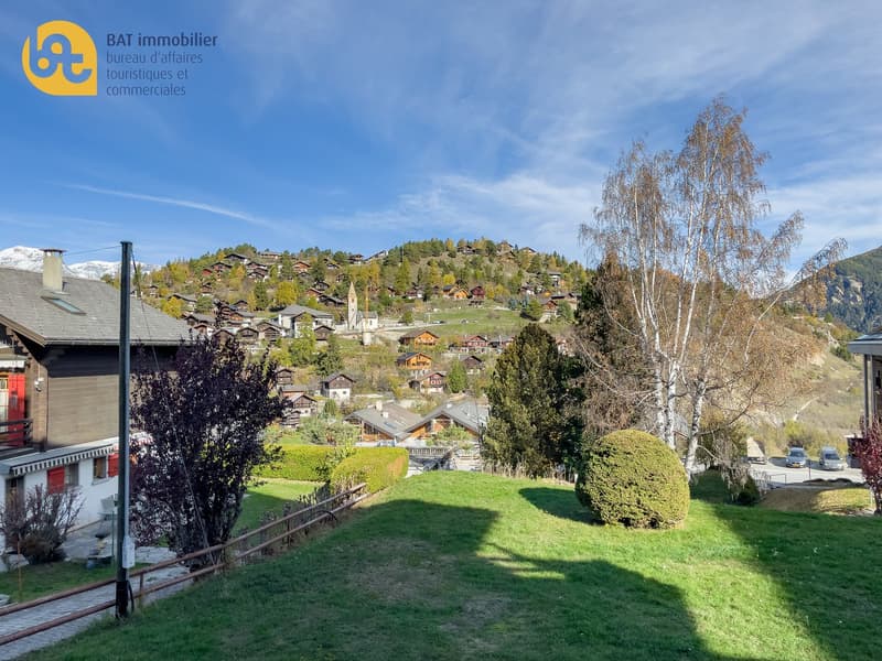 Scenic view of a mountain village with traditional Swiss-style houses, surrounded by lush green trees and vegetation. The village is situated on a hillside with a blue sky and clouds overhead. There is a grassy area in the foreground with a trimmed bush.