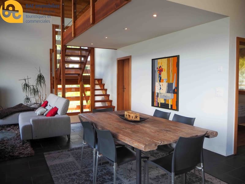 Open-concept living and dining area with wooden staircase, large wooden dining table, black chairs, gray sofa, artwork on the wall, and a fireplace