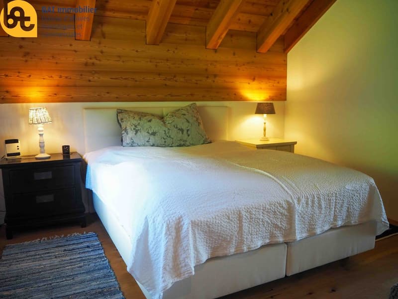 Cozy bedroom with wooden ceiling beams, a large bed with a white comforter, a side table with a lamp, and a rug on the floor.