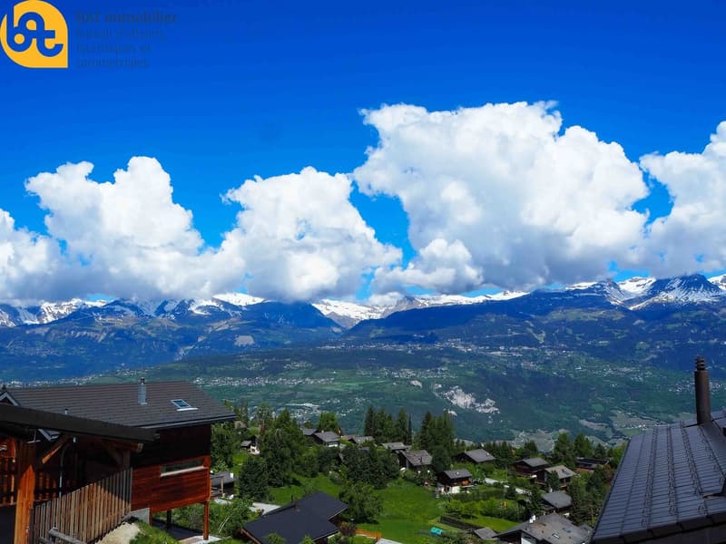 Scenic view of a mountainous landscape with snow-capped peaks, surrounded by lush green forests and a small village nestled in the valley below. The sky is filled with fluffy white clouds against a vibrant blue backdrop.