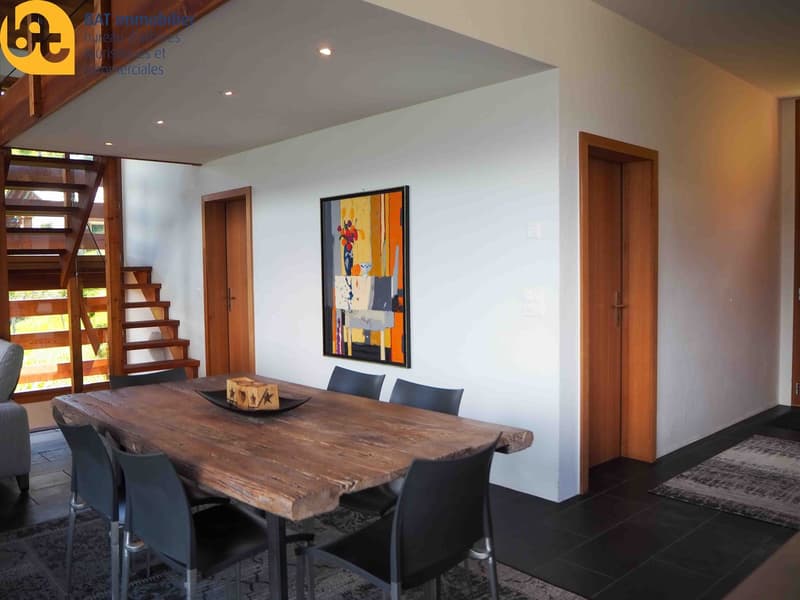 Spacious dining area with a large wooden dining table, black chairs, and a framed abstract artwork on the wall. The room has white walls, wooden stairs, and a fireplace visible.