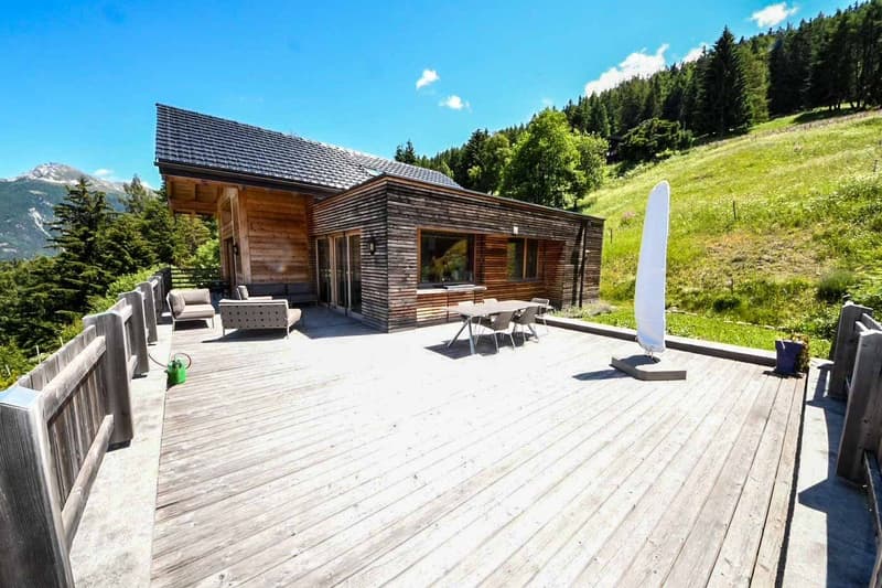 Two-story wooden cabin with a large deck overlooking a grassy hill and forested mountains in the background. The cabin has a slanted roof and is surrounded by a wooden fence. The deck features outdoor furniture including a sofa and chairs.