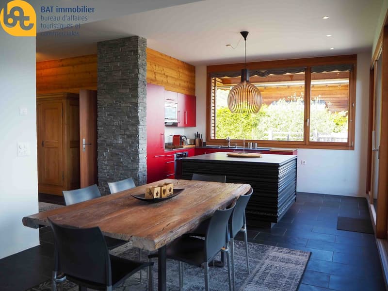 Open-concept kitchen and dining area with wooden walls, stone fireplace, red kitchen cabinets, large windows, wooden dining table, and modern pendant lighting
