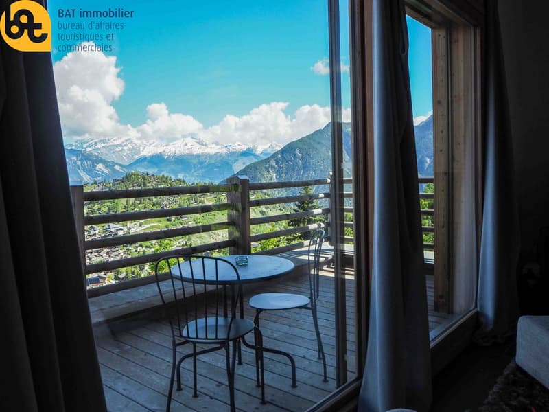 Wooden balcony with a table and chairs, overlooking a scenic mountain landscape with snow-capped peaks and lush greenery.