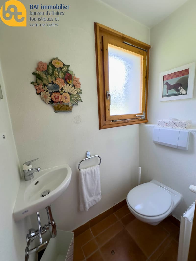 The image shows a bathroom with a sink, toilet, and a decorative floral wall hanging. The bathroom has a wooden framed window and tile flooring.
