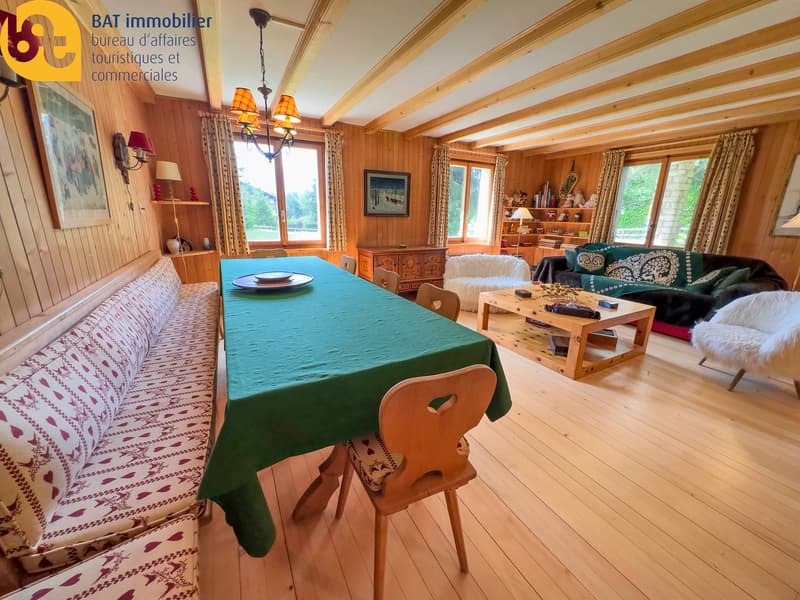 Cozy wooden interior with beamed ceiling, large windows, dining table with green tablecloth, wooden chairs, sofa, shelves, and a fireplace