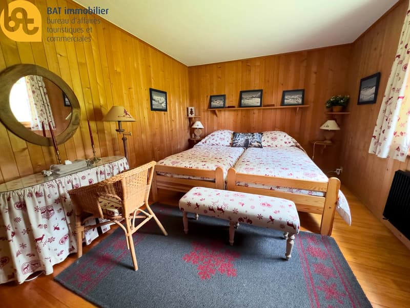 Cozy wooden bedroom with a double bed, nightstands, lamps, and a floral patterned bedding. The room has wooden paneled walls and a round mirror. There are framed artwork and shelves on the walls. A wicker chair and a patterned rug are also present in the