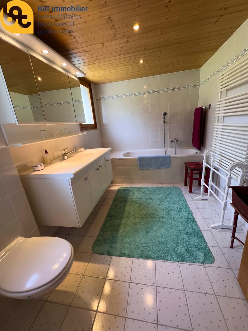 Spacious bathroom with tiled floor, vanity unit with sink, bathtub, and a green bath mat. The ceiling is made of wood paneling and the walls are tiled with a decorative border.