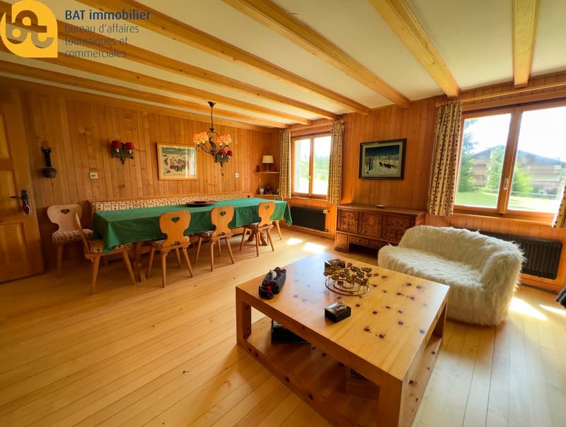 Spacious living room with wooden walls and ceiling, large windows, wooden furniture including a dining table with chairs, a sofa, and a coffee table with decorative items. The room also features a fireplace and a chandelier.