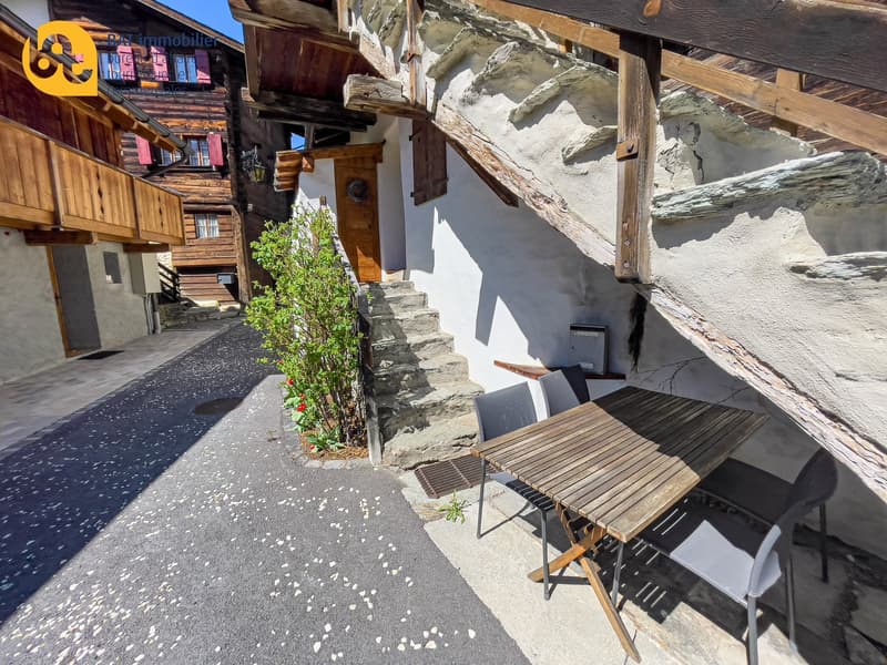 Outdoor seating area with wooden table and chairs, surrounded by stone walls and steps, with a small plant growing on the side