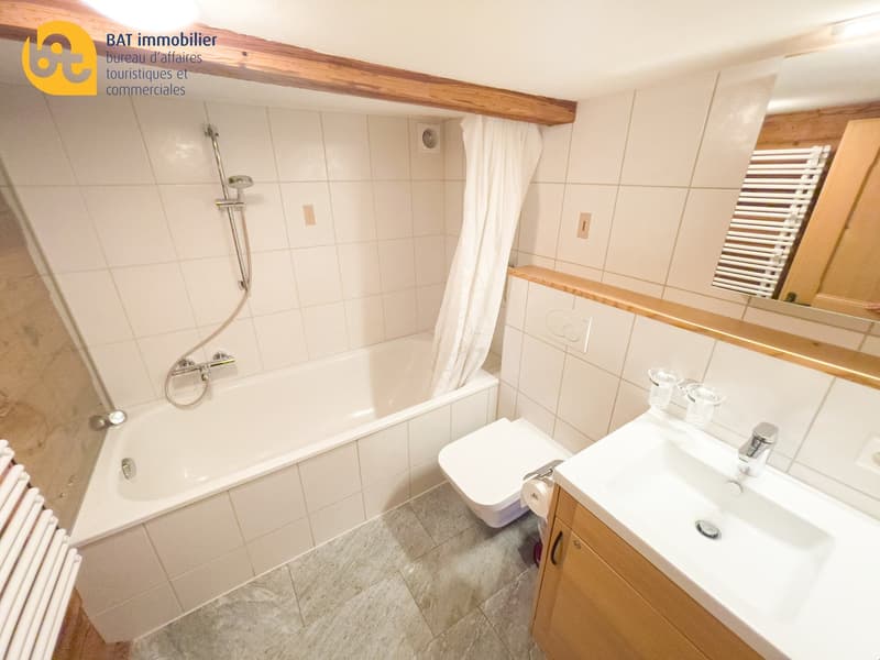 Tiled bathroom with bathtub, shower curtain, vanity with sink, and wooden shelves. Private washing machine is visible.