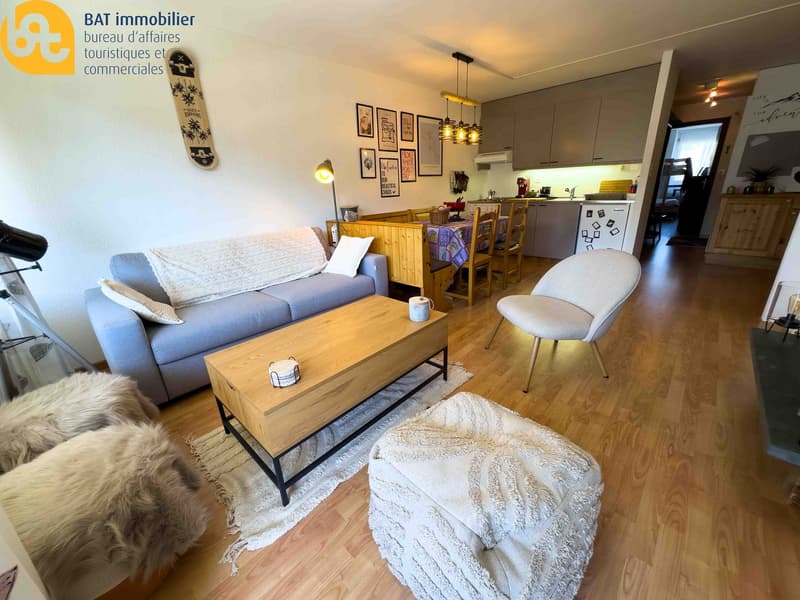 Open-plan living room and kitchen, hardwood floors, gray sofa, wooden coffee table, dining table with chairs, wall decor, snowboard on the wall
