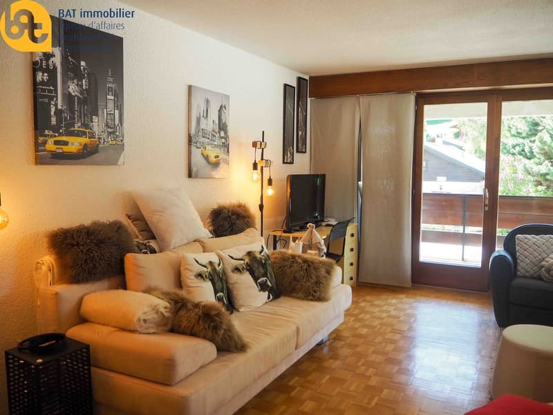 Cozy living room with large windows, hardwood floors, beige sofa with fur cushions, wall art depicting New York City, and a balcony or terrace visible through the sliding glass doors.