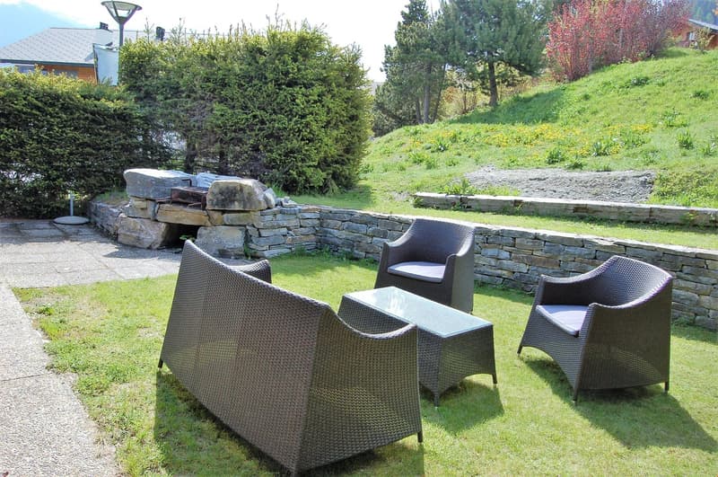 The image shows an outdoor seating area with wicker furniture, including chairs and a coffee table, set on a grassy area. There is a stone fireplace visible in the background, and the area is surrounded by lush greenery and trees.