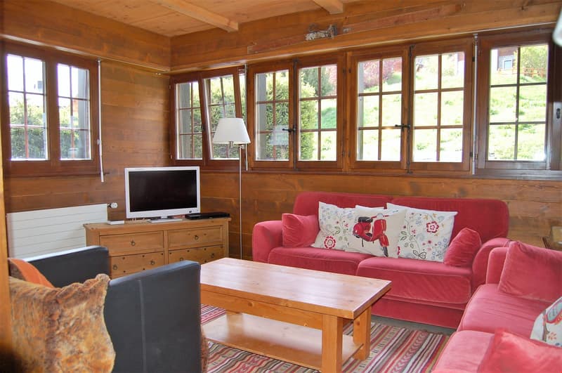 Cozy wooden cabin-style living room with large windows overlooking the outdoors, a red sofa, wooden coffee table, and a TV stand with a computer monitor.