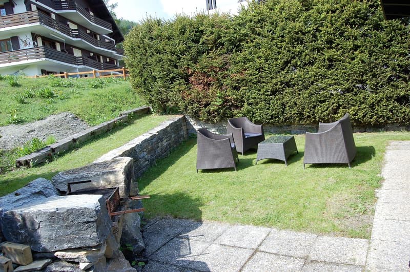 Outdoor seating area with wicker furniture, surrounded by lush greenery and a stone wall. The building in the background appears to be a hotel or resort.