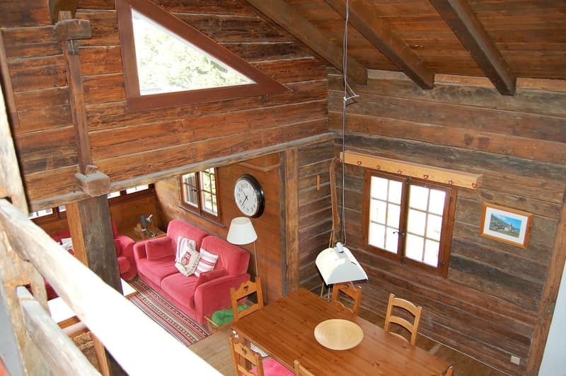 Rustic wooden cabin interior with exposed wooden beams and walls, a red sofa, a wooden dining table with chairs, and a clock on the wall. There is a fireplace visible in the living room area.