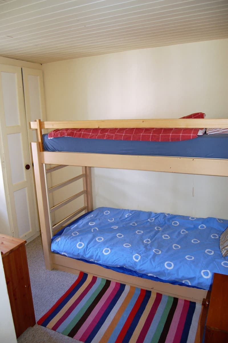 The image shows a bedroom with a bunk bed. The top bunk has a red and white patterned bedding, while the bottom bunk has blue and white patterned bedding. The room has a wooden ceiling with white painted walls. There is a wooden dresser or cabinet visible