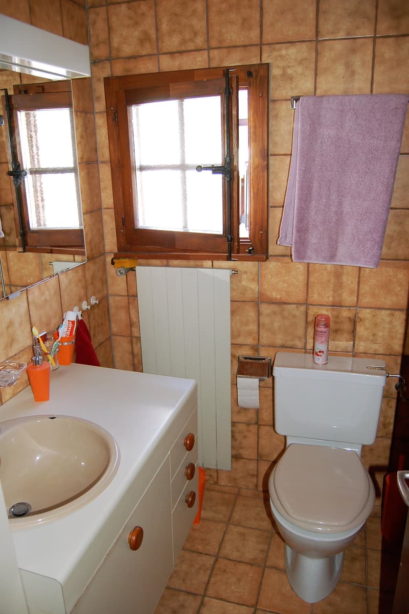 Bathroom with tile walls, wooden window frame, vanity sink, toilet, and a towel hanging on the wall.