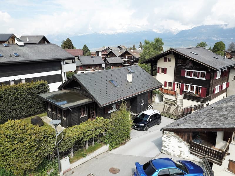 The image shows a picturesque village nestled in the mountains, with traditional Swiss-style wooden houses with red-tiled roofs. The buildings are surrounded by lush greenery, including trees and shrubs. There are several cars parked on the streets, and t