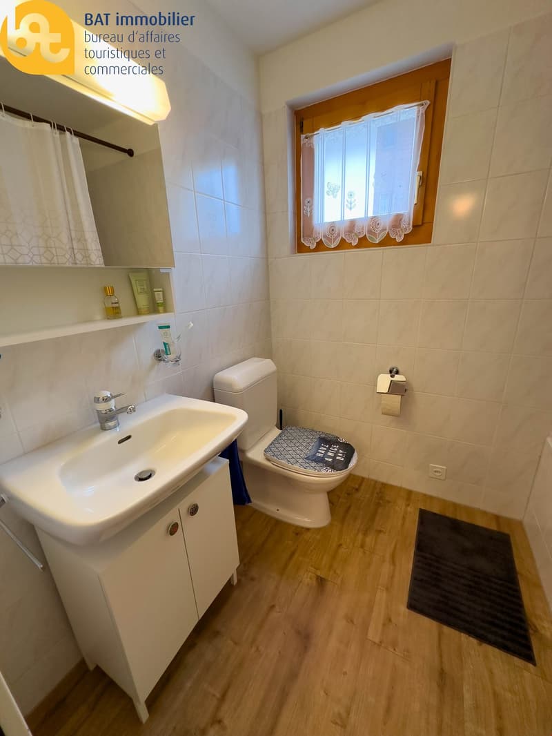 Bathroom with white tiles, wooden floor, vanity sink, toilet, and a window with lace curtains