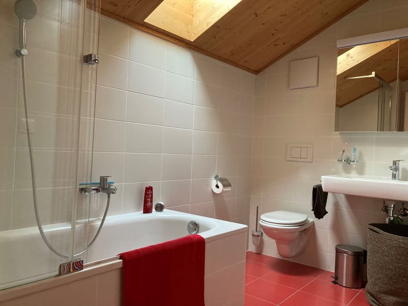 white tiled walls, white bathtub with glass door, white toilet, white sink, mirror with shelf, trash bin, trash can, red tiles on the floor, showerhead, shower curtain
