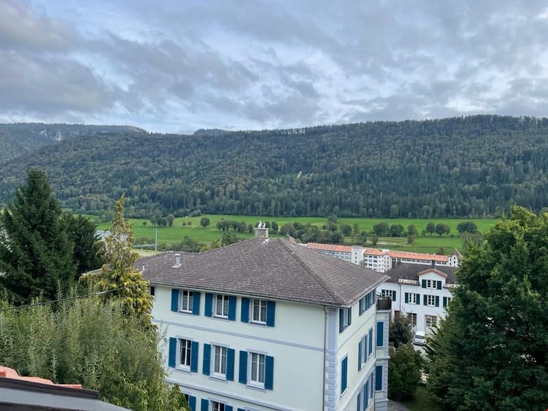multistory building, white walls, blue shutters, brown roof, green valley view in the background