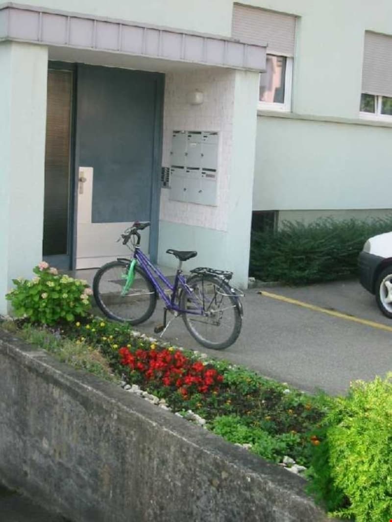 Building entrance with a bicycle parked in front, a car parked in the parking lot, and a flower bed on the left side.